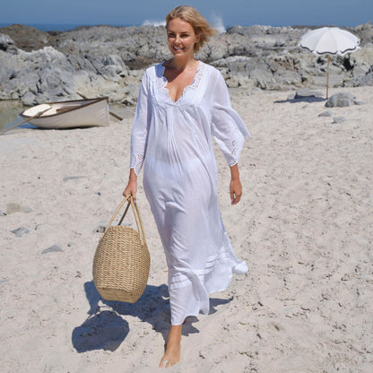 Woman in white maxi kaftan with embroidery detail at neckline and sleeve edges, carrying grass beach bag