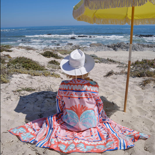 Back view of woman sitting on beach wearing relaxed fit, pink palm print beach robe